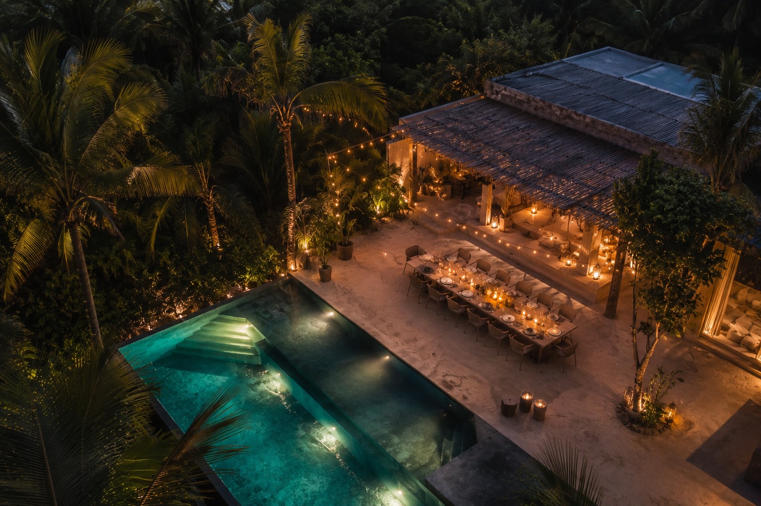 Aerial twilight view of a private Tulum villa with infinity pool, palm trees, candle-lit dinner table set for ten, and string lights overhead